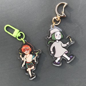May include: Two colorful keychains featuring cartoon characters. One keychain has a green-haired character holding a candle, and the other keychain has a red-haired character holding a cocktail.