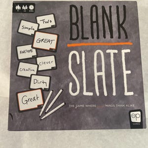 Puede incluir: Una caja de juego de mesa titulada "BLANK SLATE" con texto blanco y una l&iacute;nea naranja. La caja presenta peque&ntilde;as pizarras con palabras como "Simple" y "Great", junto con marcadores blancos. El juego es de "op GAMES".