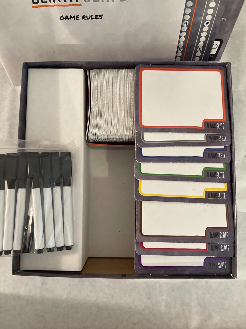 Puede incluir: Caja de juego abierta con componentes. Incluye una pila de tarjetas en blanco con bordes de colores, un juego de marcadores negros y un tablero de juego. Las tarjetas est&aacute;n etiquetadas como "BLANK SLATE". La tapa de la caja dice "GAME RULES".