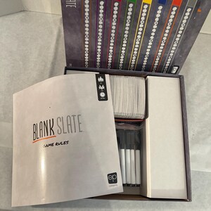 Puede incluir: El juego de mesa "Blank Slate" se muestra abierto, revelando los componentes del juego. La caja presenta el t&iacute;tulo del juego y marcadores coloridos. En el interior, hay cartas, marcadores y un libro de reglas. El juego est&aacute; dise&ntilde;ado para varios jugadores.