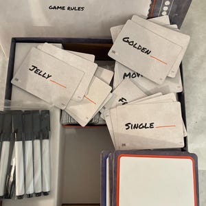 Puede incluir: Un juego de mesa con las palabras "BLANK SLATE" y "GAME RULES" impresas en la tapa. Dentro hay cartas con palabras como "JELLY", "GOLDEN" y "SINGLE", junto con cartas en blanco y rotuladores negros.