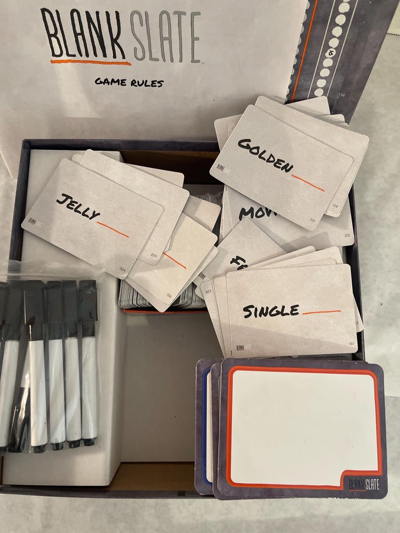 Puede incluir: Un juego de mesa con las palabras "BLANK SLATE" y "GAME RULES" impresas en la tapa. Dentro hay cartas con palabras como "JELLY", "GOLDEN" y "SINGLE", junto con cartas en blanco y rotuladores negros.