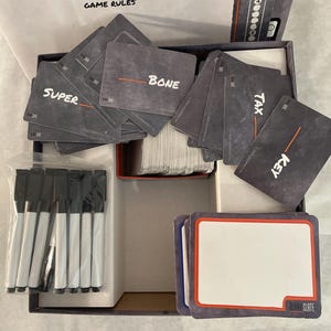 Puede incluir: Una caja de juego de mesa abierta con las palabras "BLANK SLATE" y "GAME RULES" visibles. La caja contiene cartas con palabras como "SUPER", "BONE", "TAX" y "KEY". Tambi&eacute;n se incluyen varios rotuladores de borrado en seco negros y tarjetas en blanco.