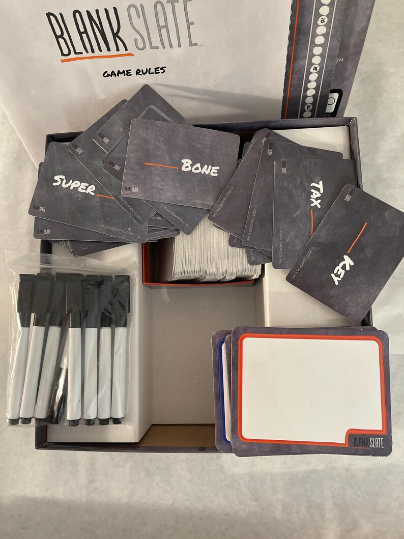 Puede incluir: Una caja de juego de mesa abierta con las palabras "BLANK SLATE" y "GAME RULES" visibles. La caja contiene cartas con palabras como "SUPER", "BONE", "TAX" y "KEY". Tambi&eacute;n se incluyen varios rotuladores de borrado en seco negros y tarjetas en blanco.