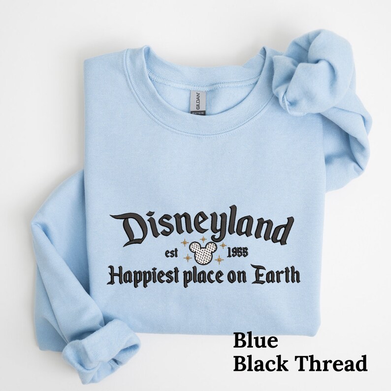 May include: Light blue sweatshirt with black embroidered text that reads "Disneyland est 1955 Happiest place on Earth" with a black Mickey Mouse head graphic. The text is in a vintage style font.