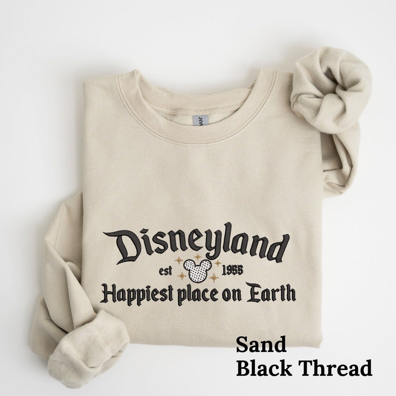 May include: A sand colored sweatshirt with black embroidery that reads "Disneyland est 1955 Happiest place on Earth". The embroidery includes a black and white Mickey Mouse head with gold stars.