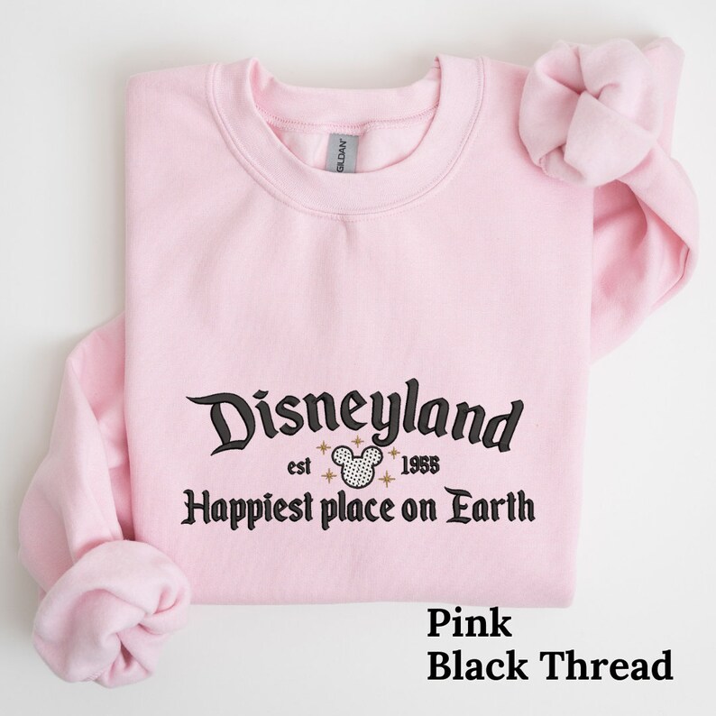 May include: Pink sweatshirt with black text that reads "Disneyland est. 1955 Happiest place on Earth" with a black and white Mickey Mouse head graphic.