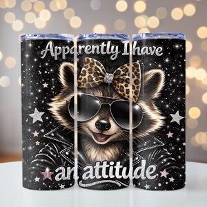 May include: A set of three black stainless steel tumblers with a design featuring a raccoon wearing sunglasses, a leopard print bow, and a black leather jacket. The text on the tumbler reads "Apparently I have an attitude."