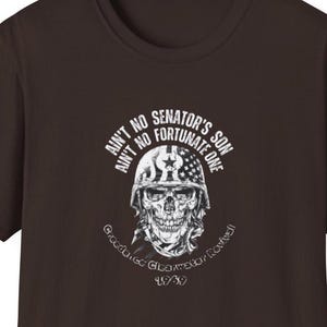 May include: A brown t-shirt featuring a white graphic of a skull wearing a military helmet with a star and stripes design. The text reads "Ain't No Senator's Son Ain't No Fortunate One" and "Creedence Clearwater Revival 1969."
