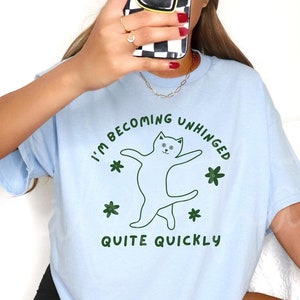 May include: A light blue t-shirt with a graphic of a cat dancing and the text "I'm becoming unhinged quite quickly".