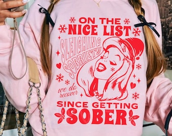 May include: Pink sweatshirt with a red graphic design featuring a woman in a Santa hat with the text "On the Nice List Sleighing Sobriety We do recover Since Getting Sober".