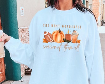 May include: A light blue sweatshirt with the text "The most wonderful season of them all" and a graphic of a pumpkin, a football, a candle, and a coffee cup.