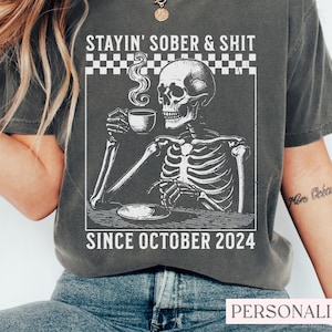 May include: Dark gray t-shirt with a graphic of a skeleton drinking coffee, with the text "STAYIN' SOBER & SHIT SINCE OCTOBER 2024". The design is in white, with a checkered border at the top. The shirt is a relaxed fit.