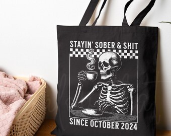 May include: Black tote bag with a skeleton illustration drinking from a cup, and the text "STAYIN' SOBER & SHIT SINCE OCTOBER 2024". The bag has black handles and a checkered border.
