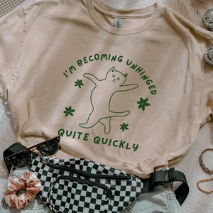 May include: A beige t-shirt with a green graphic of a cat with the text "I'm becoming unhinged quite quickly".