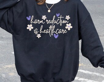 May include: Black crew neck sweatshirt featuring the phrase "harm reduction is healthcare" in white, with purple heart and white flower accents. A casual, comfortable garment.