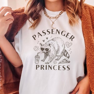 May include: White t-shirt with a graphic of a raccoon wearing sunglasses and holding a drink. The text "Passenger Princess" is printed above and below the raccoon.