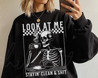 May include: Black sweatshirt featuring a skeleton drinking coffee, with the text "LOOK AT ME" above and "STAYIN' CLEAN & SHIT" below. The graphic is in black and white. The sweatshirt has long sleeves.