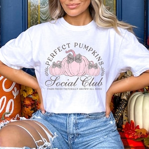 May include: White t-shirt with a graphic of three pink pumpkins with black and white bows. The text "Perfect Pumpkins Social Club" is written in brown with the words "Farm Fresh - Naturally Grown - All Sizes" underneath.