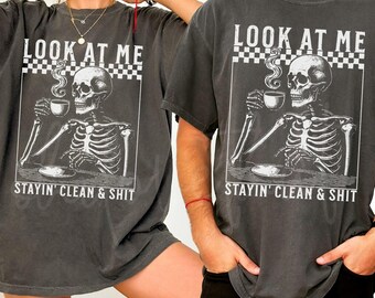 May include: Two grey t-shirts featuring a skeleton graphic holding a cup of coffee. The text on the shirts reads "Look at me Stayin' Clean & Shit".