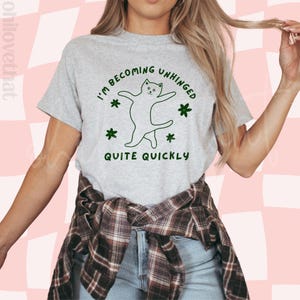 May include: A grey t-shirt with the text "I'm becoming unhinged quite quickly" and a green line drawing of a cat dancing. The cat is surrounded by green flowers.