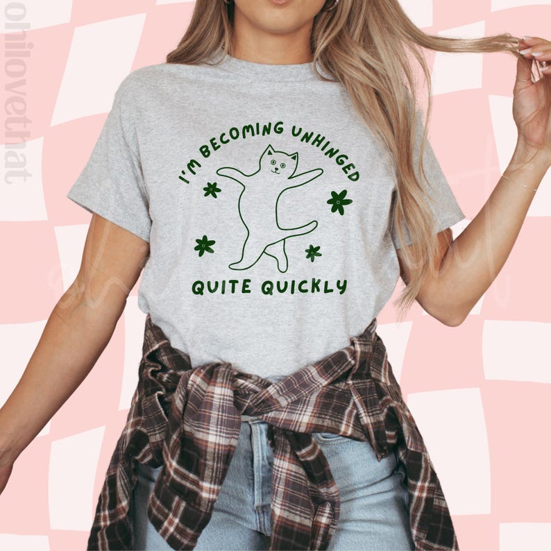 May include: A grey t-shirt with the text "I'm becoming unhinged quite quickly" and a green line drawing of a cat dancing. The cat is surrounded by green flowers.