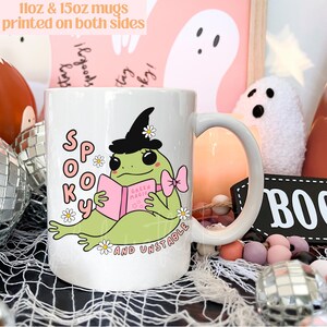 May include: White ceramic mug with a green frog wearing a witch hat and reading a book that says "Green Magic". The frog is surrounded by white daisies and the text "Spooky and Unstable" is printed below the frog.