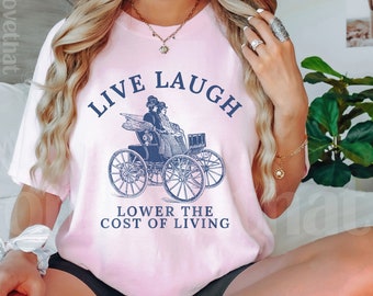 May include: A light pink t-shirt with the words "LIVE LAUGH" above a vintage carriage illustration. Below the carriage, the text reads "LOWER THE COST OF LIVING". The shirt is a soft, casual style.