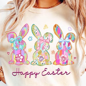 May include: A white t-shirt with a graphic of three pastel colored bunnies with bows and the text "Happy Easter" printed on the front.
