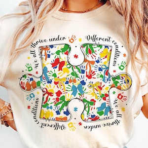 May include: A white t-shirt with a colorful puzzle piece design. The puzzle piece is filled with handprints and bows. The text around the puzzle piece reads "We all thrive under different conditions".
