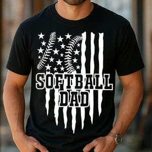 Op de afbeelding: Zwart T-shirt met een witte afbeelding van een softbal en sterren, die lijken op de Amerikaanse vlag, met de tekst "Softball Dad" eronder.