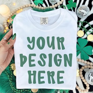 May include: A white t-shirt with the text "YOUR DESIGN HERE" in green block letters. The shirt is a Comfort Colors brand, size M-M-M. The shirt is surrounded by St. Patrick's Day themed decorations, including shamrocks and gold coins.