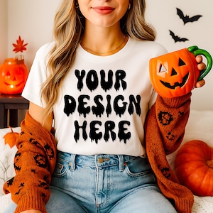 May include: White t-shirt with the text "YOUR DESIGN HERE" in black, worn with blue jeans. A person is holding an orange pumpkin-shaped mug with a green handle. The person is wearing an orange sweater. Halloween theme.