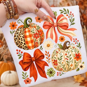 May include: A white fabric surface featuring a printed design of two pumpkins and two bows. One pumpkin has leopard print, plaid, and floral sections. The other is cream with floral vines. Two large orange and striped bows are surrounded by small flowers.