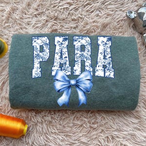 May include: A teal sweatshirt with the word "PARA" in large white letters filled with a blue floral pattern. A blue bow is centered below the text. The sweatshirt is on a fluffy, light-colored surface.