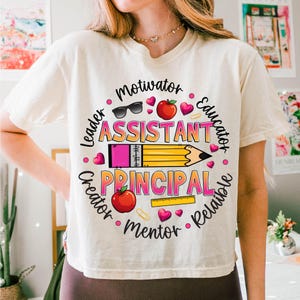 May include: Cream-colored t-shirt featuring a colorful graphic design. The design includes the words "Assistant Principal" with surrounding words like "Motivator" and "Educator". The design also includes a pencil, hearts, and apples.