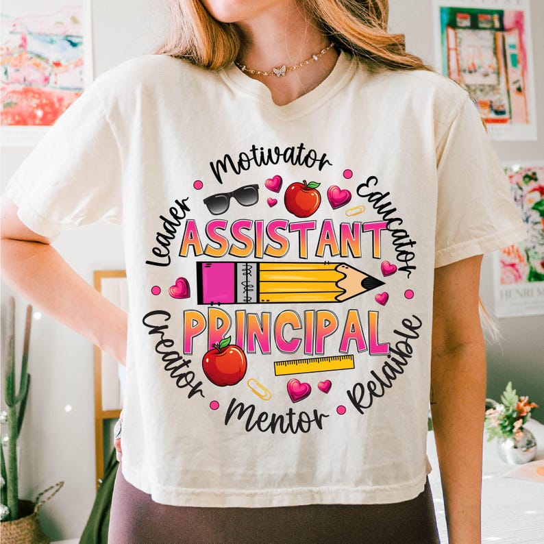 May include: Cream-colored t-shirt featuring a colorful graphic design. The design includes the words "Assistant Principal" with surrounding words like "Motivator" and "Educator". The design also includes a pencil, hearts, and apples.