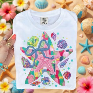 May include: White t-shirt featuring a colorful patchwork star design with a pink starfish and blue bow. The shirt is adorned with embroidered seashells in various colors. The tag reads "COMFORT COLORS" and "M-M-M".