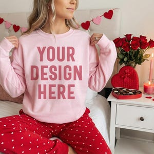 May include: A light pink sweatshirt with the text "YOUR DESIGN HERE" in a darker pink font. The person is wearing red pajama pants with white hearts. A Valentine's Day theme is present with heart decorations and a heart-shaped box of chocolates.