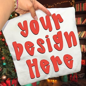 May include: White pillow with the text "YOUR DESIGN HERE" in red, outlined letters. The pillow is being held up by a hand. The background includes a Christmas tree and a bookshelf.