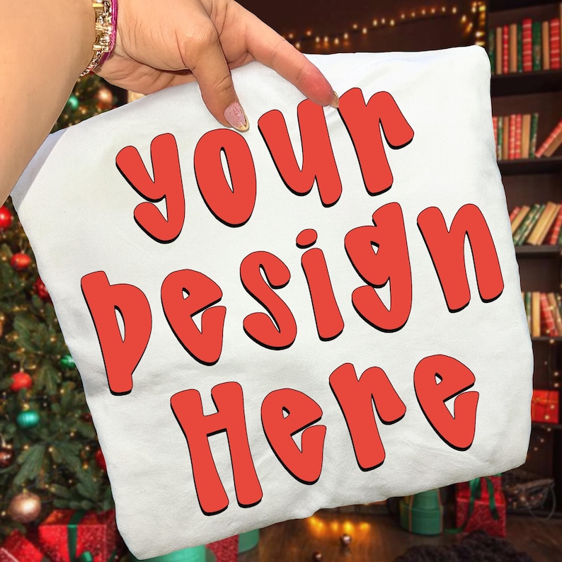 May include: White pillow with the text "YOUR DESIGN HERE" in red, outlined letters. The pillow is being held up by a hand. The background includes a Christmas tree and a bookshelf.