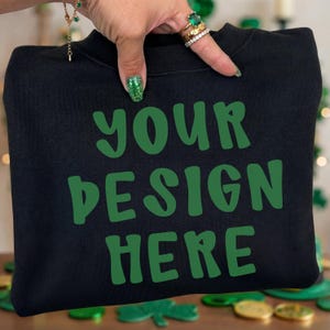 Puede incluir: Sudadera negra con el texto "YOUR DESIGN HERE" en verde, perfecta para diseños personalizados. La sudadera es sostenida por una mano con esmalte de uñas verde brillante y anillos. El fondo tiene tréboles verdes.