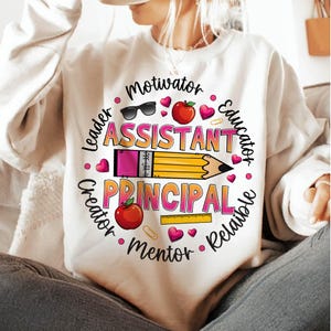 May include: White sweatshirt with a colorful graphic design for an assistant principal. The design includes the words "Assistant Principal" and other words like "Motivator" and "Educator" around a pencil and other school-themed elements.