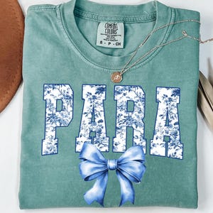 May include: A seafoam green t-shirt with the word "PARA" in white block letters filled with a blue floral pattern. A large blue bow is below the text. A gold necklace with a pendant is on the shirt.