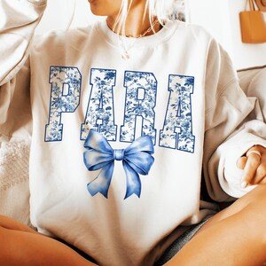 May include: White sweatshirt with the word "PARA" in blue floral print letters. A blue bow is centered below the text. The sweatshirt is a casual style.