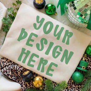Może przedstawiać: Kremowa bluza z napisem "YOUR DESIGN HERE" w kolorze zielonym. Bluza otoczona jest dekoracjami na Dzień Świętego Patryka, w tym zielonymi ozdobami, garnkiem złota i kapeluszem krasnoludka.
