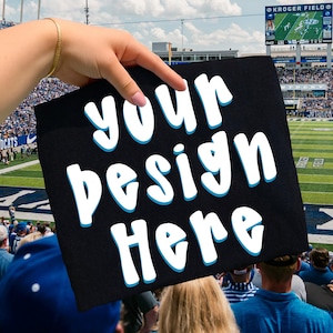 May include: A black sign with the words "your design Here" in white, outlined in blue. The sign is held up in front of a football stadium filled with people. The sky is cloudy.