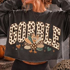 May include: A charcoal gray long-sleeve sweatshirt with a raw-edge hem. The front features the word GOBBLE in large block letters with a leopard print pattern. Below the text is a cartoon turkey with patterned feathers and a small bow.