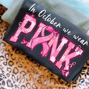 May include: A dark gray t-shirt with the words "In October we wear PINK" in white script. The word "PINK" is in large pink letters with a patchwork design and pink ribbon accents, and pink butterflies.