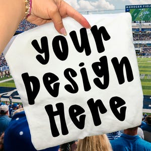 May include: A white pillow with the words "your design here" in bold black letters. The pillow is held up in front of a football stadium. The background shows a football field and spectators.